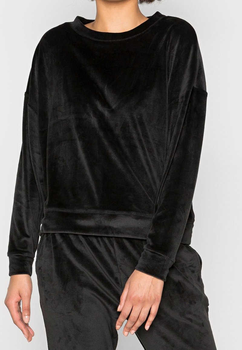 Femme portant un haut en velours noir à manches longues et un pantalon assorti en velours noir, debout avec les bras détendus le long du corps sur un fond uni.