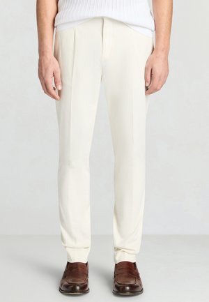 Mann, der maßgeschneiderte weiße Anzughosen und braune Lederloafers trägt, steht vor einem schlichten hellen Hintergrund.