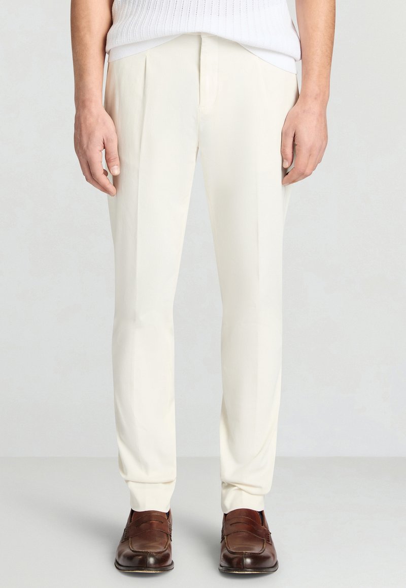 Mann, der maßgeschneiderte weiße Anzughosen und braune Lederloafers trägt, steht vor einem schlichten hellen Hintergrund.