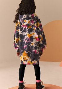 Bambina con lunghi capelli scuri che indossa un vestito ampio con cappuccio in tinta unita nero, giallo e rosa, leggings neri, calze rosa e scarpe alte nere.