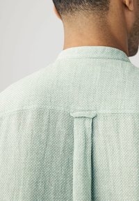 Camisa texturizada verde claro con cuello de punta, que presenta un pliegue central en la parte posterior y un lazo para colgar. La tela muestra un pequeño patrón tejido.