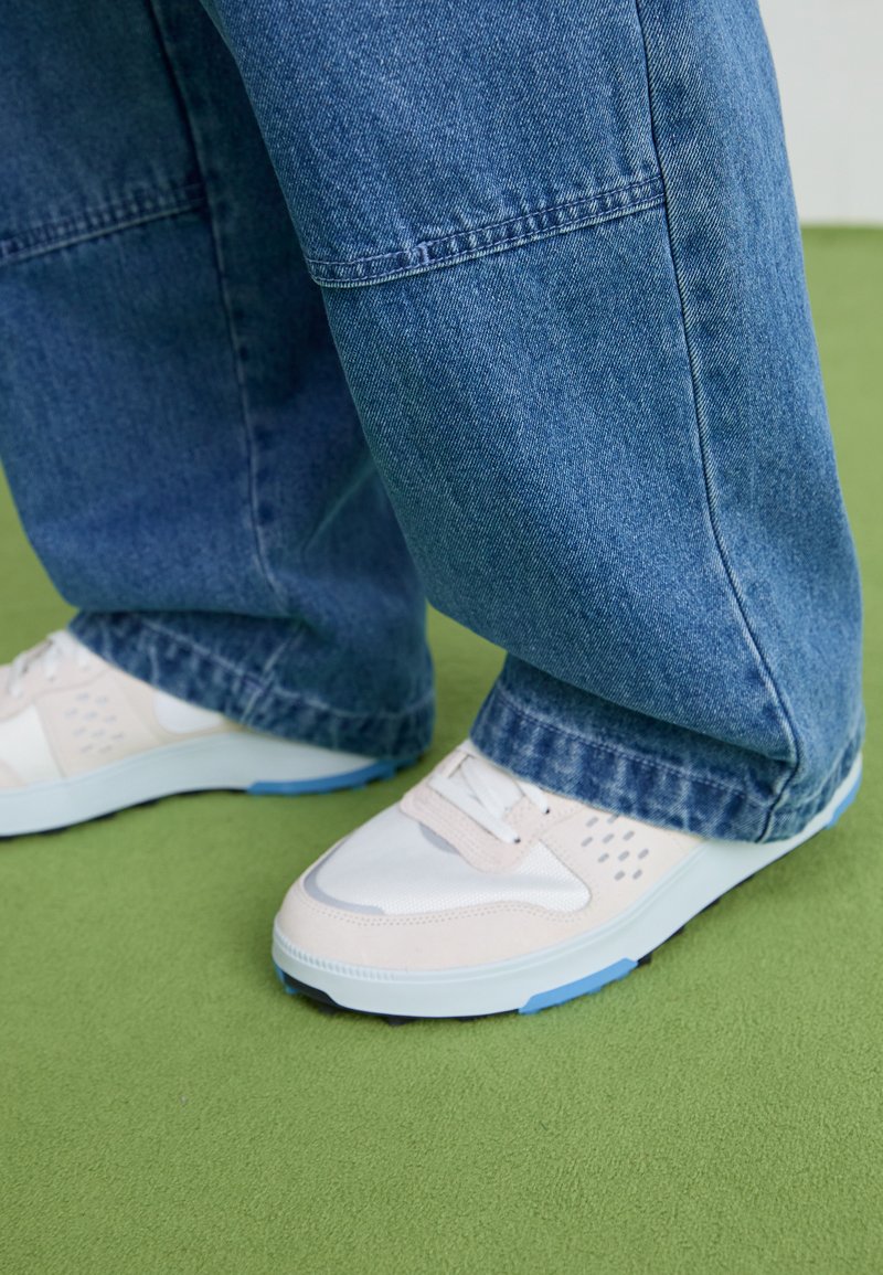Zapatillas blancas con detalles en rosa claro y suelas azules, combinadas con pantalones de denim azul holgados. Textura suave y un diseño minimalista.