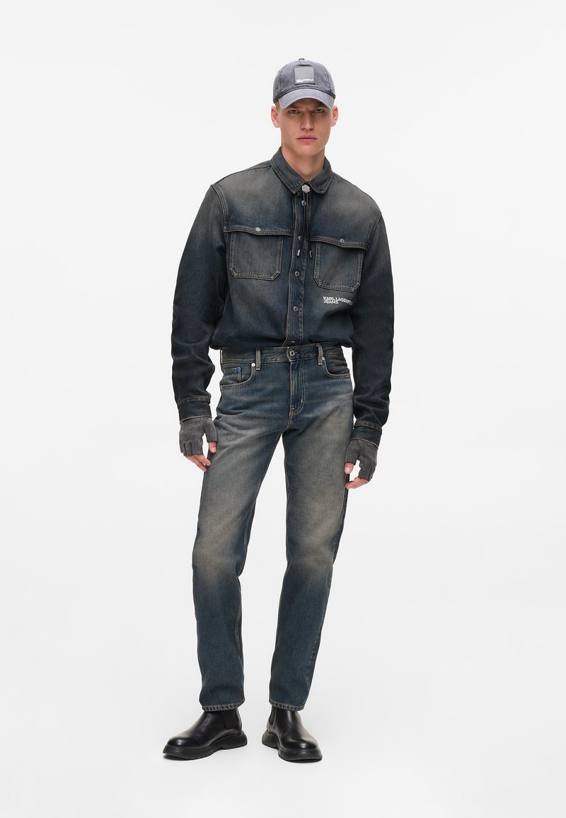 Denim jacket with two chest pockets, dark blue color, worn over faded jeans, paired with a gray cap and black boots. Notable fingerless gloves.