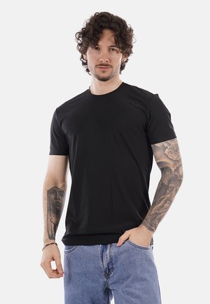 Uomo con capelli ricci che indossa una maglietta nera e jeans blu, mostra tatuaggi su entrambe le braccia, in piedi davanti a uno sfondo semplice.