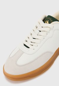 White leather sneaker with beige suede accents, flat laces, and a gum rubber sole. Features a green logo tag at the heel.