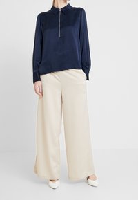 Camicia a maniche lunghe navy con zip a placket, abbinata a pantaloni a gamba larga color beige chiaro, caratterizzati da texture morbide e una silhouette rilassata.