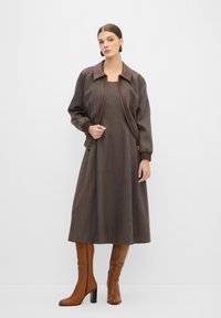 Veste aviateur marron avec un col sombre brillant, assortie à une robe marron texturée et des bottes en cuir marron montantes avec un talon haut.