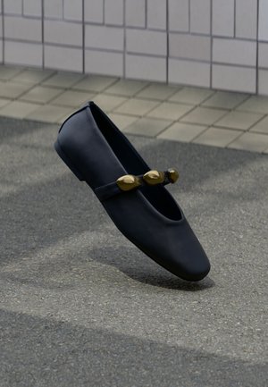 Fekete bőr lapos cipő, pánttal, amelyen három nagy arany díszgyöngy található, egy burkolt talaj és csempézett fal előterében.