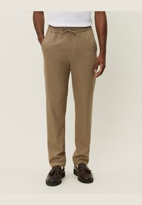 Pantalones de color marrón claro, con textura, cinturilla elástica y cordón. Cuentan con bolsillos laterales y un ajuste holgado, combinados con zapatos oscuros.
