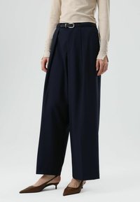 Marineblauwe wijde broek met hoge taille, plooien en een zwarte riem. Gecombineerd met een beige top en bruine pumps met spits neusje.