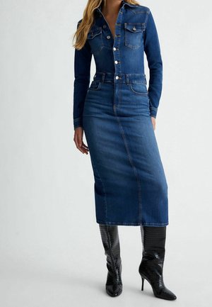 Femme portant une robe en jean bleu cintrée à boutons avec manches longues et des bottes noires texturées à talons montant jusqu'aux genoux.