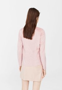 Giacca in suede rosa chiaro con maniche lunghe e design aderente, caratterizzata da cuciture sul retro. Indossata sopra una gonna beige.
