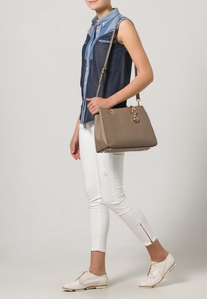 Sac à main en cuir gris avec ferrures dorées et logo, porté avec une chemise en denim bleu et un jean skinny blanc, assorti à des chaussures blanches à lacets.