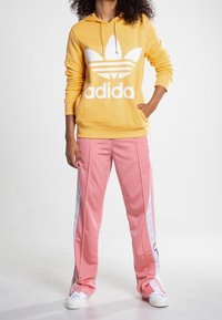 Gul huvtröja med stor vit Adidas-logga, matchad med rosa träningsbyxor med vita sidostrippar och sportiga vita sneakers.