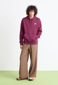 Persona con cabello rizado, vistiendo una sudadera de color burdeos, pantalones anchos de cuadros marrones y zapatillas rosa-negras, de pie sobre un suelo verde.
