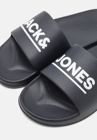Fekete csúszós szandál, texturált szintetikus anyagból, a széles pánton fehér "JACK&JONES" logóval. Lapos talp kialakítás.