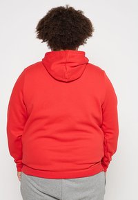 Sudadera roja hecha de un tejido suave, con un bolsillo frontal y capucha ajustable con cordón. La vista muestra la parte posterior, con una textura lisa.