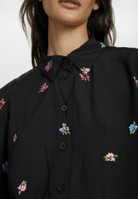 Schwarzes Hemd mit Knöpfen und floraler Bestickung in Pink, Lila und Blau. Hat einen Kragen und eine glatte Textur.