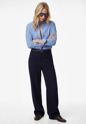 Femme blonde portant de grandes lunettes de soleil noires, une chemise rayée bleu et blanc, une ceinture noire, un pantalon large bleu marine et des chaussures marron, debout les bras croisés.
