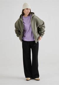 Hoodie zippé oversize de couleur vert olive, t-shirt violet clair, pantalon large noir, bonnet en tricot crème et chaussures beiges avec des accents marron.
