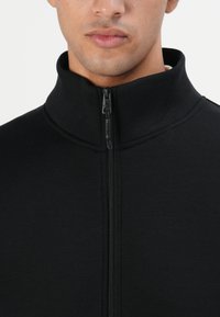 Joven vestido con una chaqueta negra de cuello alto y cremallera, mostrando el tirador de la cremallera y el área del cuello, con un fondo neutro.