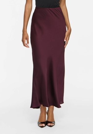 Femme portant une longue jupe en satin violet foncé associée à des talons ouverts noirs, debout devant un fond clair uni.