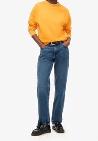 Sudadera naranja, vaqueros de mezclilla azul, cinturón negro con hebilla plateada y zapatos negros. El conjunto presenta un corte relajado y elementos de diseño clásicos.