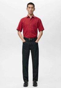 Rotes kurzärmeliges Hemd mit Knopfleiste vorne, kombiniert mit schwarzer Jeanshose und schwarzen Schuhen. Einfaches Design und passgenaue Schnittführung.