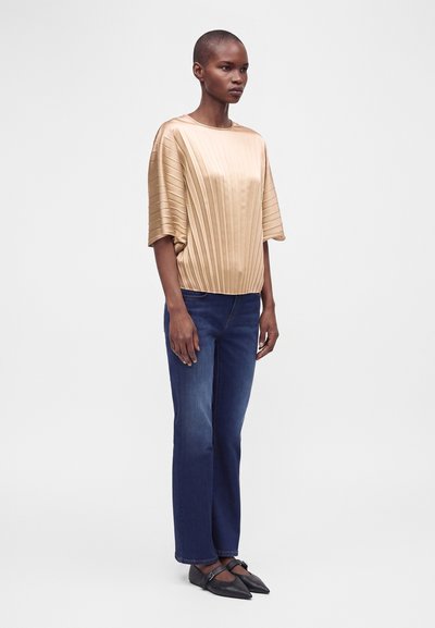 Femme debout portant un haut beige plissé à manches courtes, un jean bleu droit et des chaussures Mary Jane noires à bout pointu.