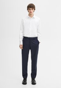 Jonge man staand, gekleed in een wit overhemd met knopen, marineblauwe broek met krijtstreep en zwarte nette schoenen, tegen een effen witte achtergrond.