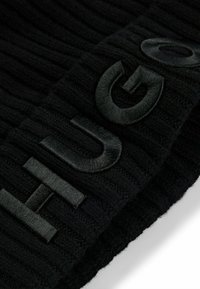 Svart strikket lue med hevet svart broderi som staver "HUGO." Stoffet har en teksturert ribbestruktur.