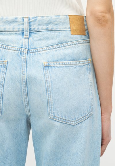 Jeans en denim bleu clair avec poches arrière et un patch de marque en cuir marron, portés par une personne portant un haut blanc côtelé.