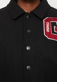 Svart ullblandad jacka med knappfront, skjortkrage och ett rött och vitt broderat märke med ordet "Originals."