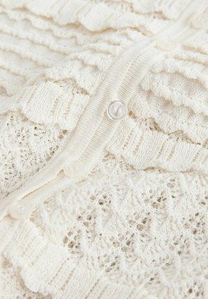 Kremasto obarvan pleten jopič z rebrasto teksturo, čipkastimi vzorci in šestimi prozornimi gumbi spredaj. Mehka, lahka tkanina.