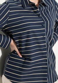 Camicia blu navy con righe orizzontali crema, apertura con bottoni e colletto. Presenta maniche lunghe e una tasca laterale con orlo curvo.