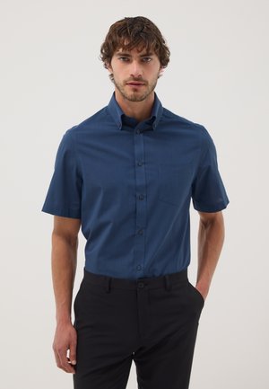 Hombre con cabello corto y rizado que lleva una camisa azul oscuro de manga corta abotonada metida dentro de pantalones negros, de pie con una mano en el bolsillo.