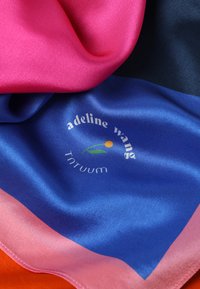 Pañuelo de seda con vibrantes tonos de rosa, azul royal y naranja, de textura suave. Logos notables y detalles de costura visibles cerca de los bordes.