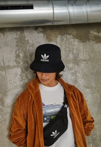 Ung person som bär en svart Adidas bucket hat och axelremsväska, brun jacka och vit t-shirt med grafik mot en betongvägg.