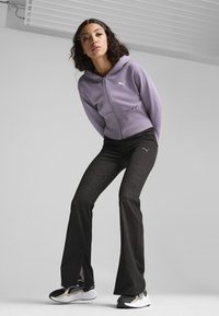 Felpa zipata viola con cappuccio e logo bianco, abbinata a pantaloni neri svasati con un motivo testurizzato e sneakers nere.