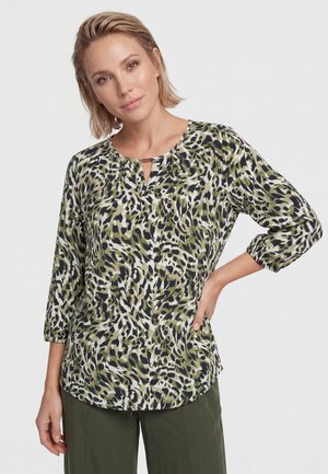 Donna con capelli biondi corti che indossa una camicetta verde, nera e bianca con stampa animalier, maniche a tre quarti e pantaloni verde scuro.