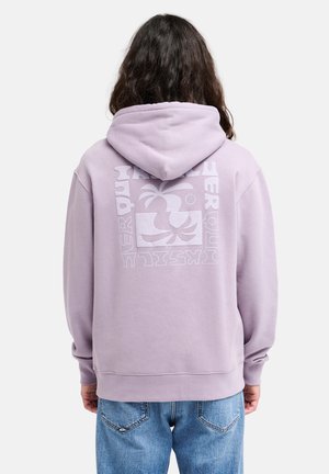 Personne aux longs cheveux portant un sweat à capuche violet clair avec un motif de palmier et un texte au dos, associée à un jean bleu.