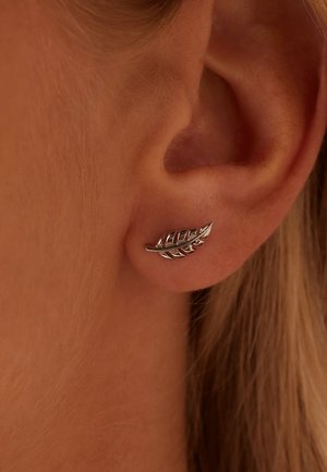 Boucle d'oreille en forme de feuille argentée avec des détails complexes, attachée au lobe de l'oreille, présentant une texture polie et un design élégant.