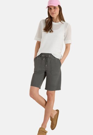 Kvinde iført en pink kasket, hvid mesh kortærmet top, mørkegrå snøreshorts og brune slip-on sko, der står med hænderne i lommerne.