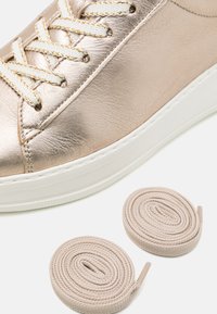 Sneaker color oro rose metallico con suola bianca, caratterizzato da lacci chiari. Due lacci beige avvolti sono posizionati accanto alla scarpa.