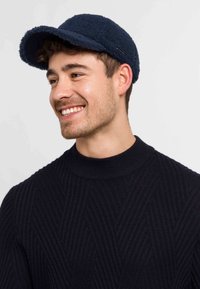 Cappello in pile blu navy con corona arrotondata e visiera curva, caratterizzato da un sottile accento metallico. Indossato con un maglione nero testurizzato.