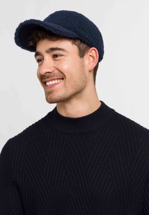 Navy blue fleece cap with a rounded crown and a curved brim, featuring a subtle metallic accent. Worn with a textured black sweater.