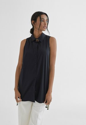Blusa negra sin mangas con cierre de botones, cuello puntiagudo y acentos de hardware decorativos. Tejido ligero y corte relajado, combinada con pantalones blancos.