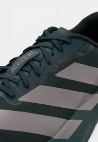 Baskets de sport vertes avec une tige texturée, des rayures grises et des lacets assortis. Présente une étiquette de marque "ADIZERO" sur la languette.