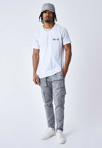 T-shirt bianco a maniche corte con logo, pantaloni cargo grigi con molte tasche e sneaker bianche. Un cappellino con design a pattern completa il look.