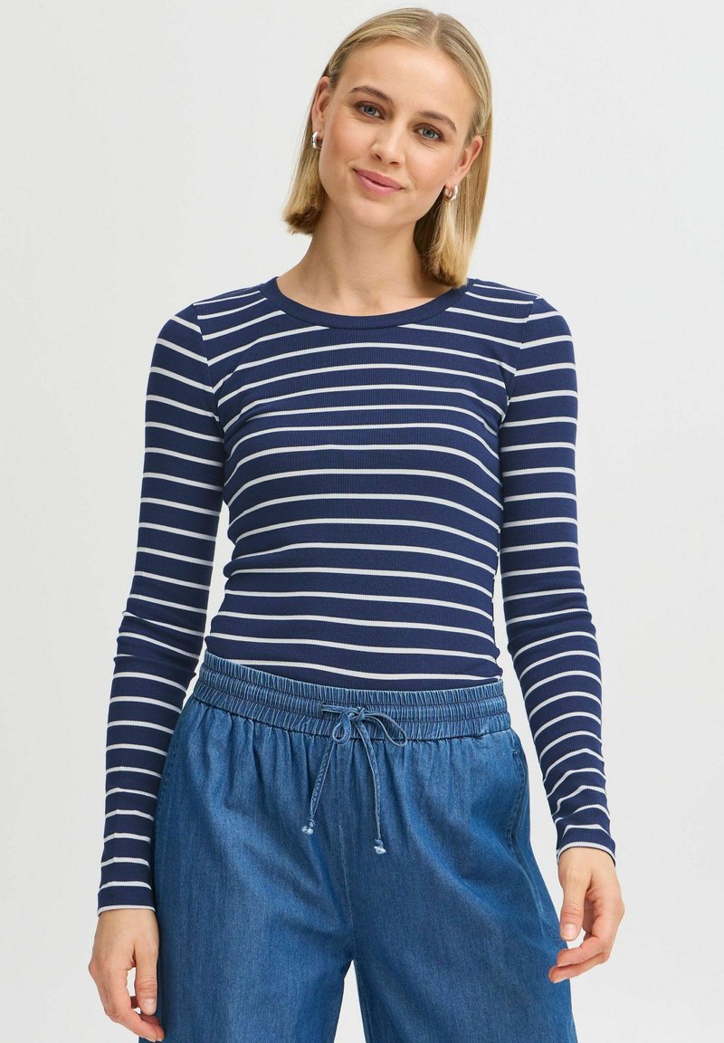 Marinebluse mit langen Ärmeln, weißen Streifen, geripptem Material und rundem Ausschnitt; kombiniert mit blauen Jogginghosen mit Kordelzug.